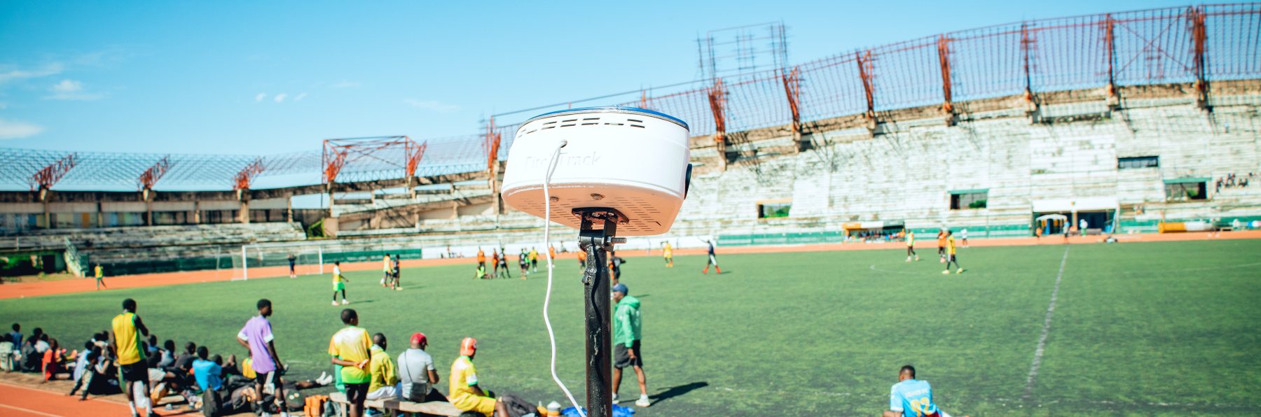 Wide view of the rig on its mast beside a full match, players on the pitch and substitutes on the touchline behind it.