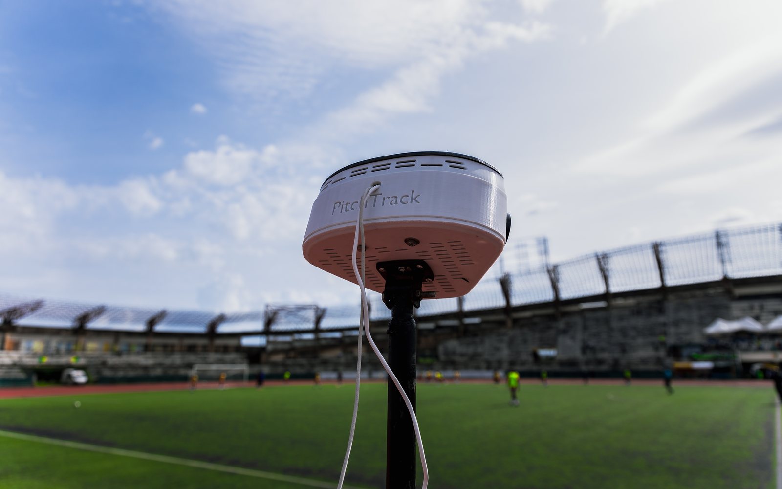 The PitchTrack rig on its mast beside the pitch, seen from below against the sky, with players and the stadium stand behind it